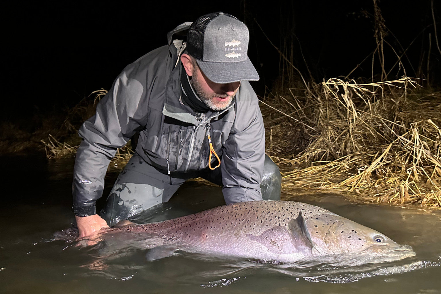 Rybár stojaci vo vode počas súmraku, oblečený v nepremokavej bunde a čiapke – funkčné rybárske oblečenie do chladného počasia.